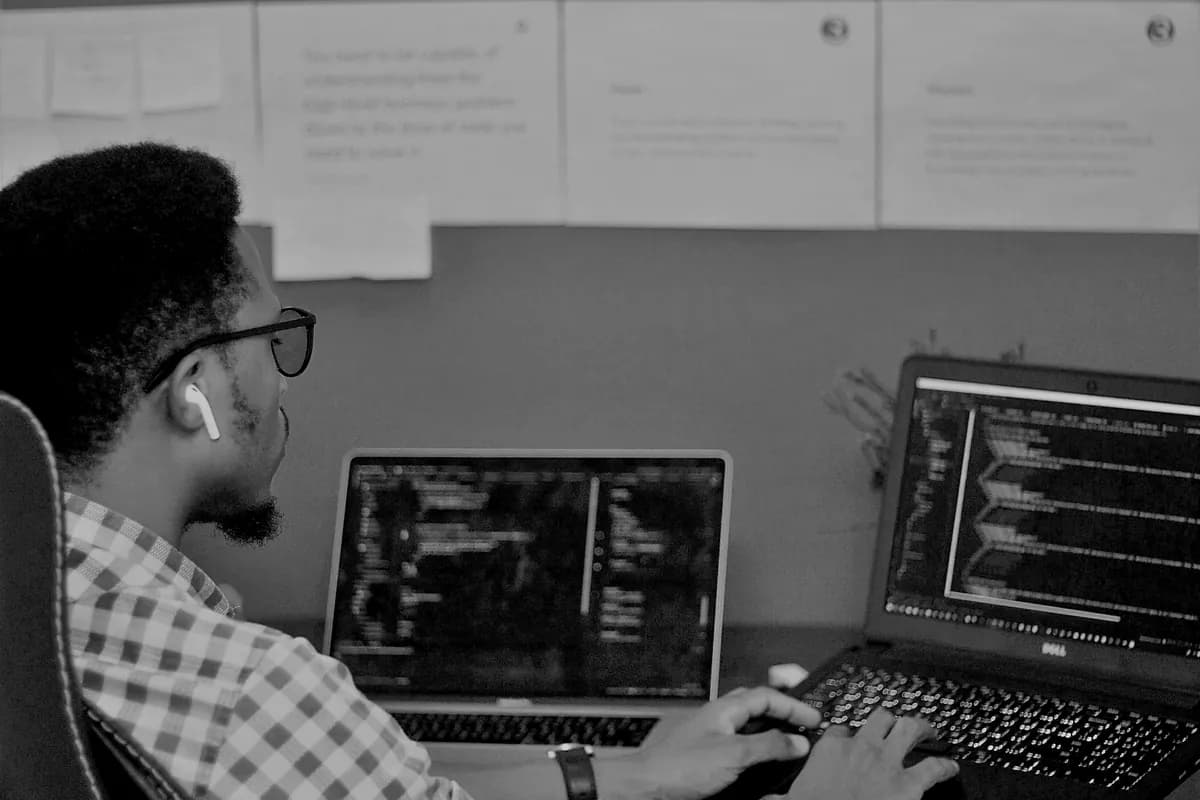 An engineer working across two laptops, with planning notes pinned to the wall above the desk.
